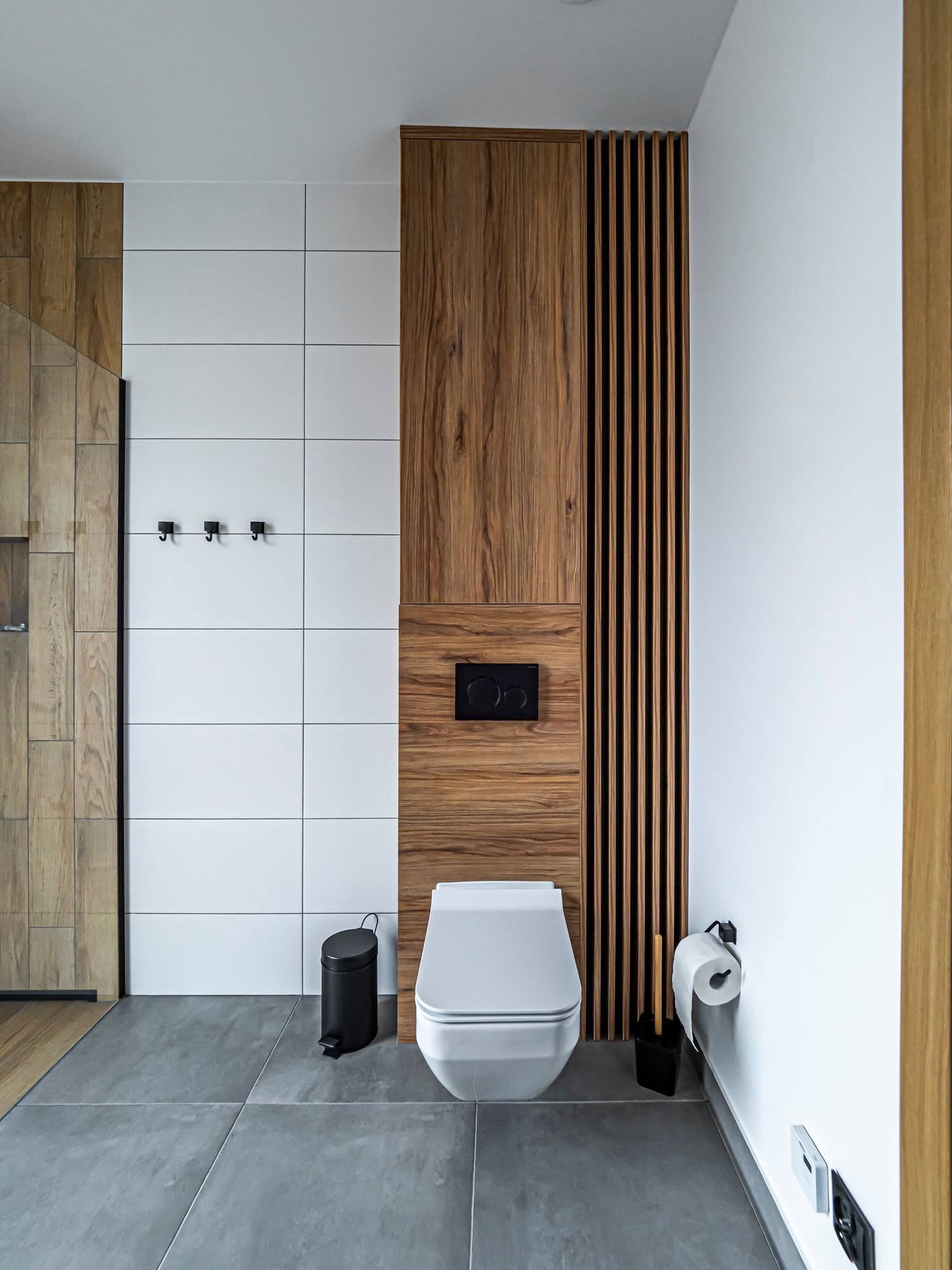 Nowoczesna łazienka z drewnianą zabudową, białymi płytkami i szarą podłogą. Uwagę zwraca drewniana ścianka z pionowymi listwami obok toalety.