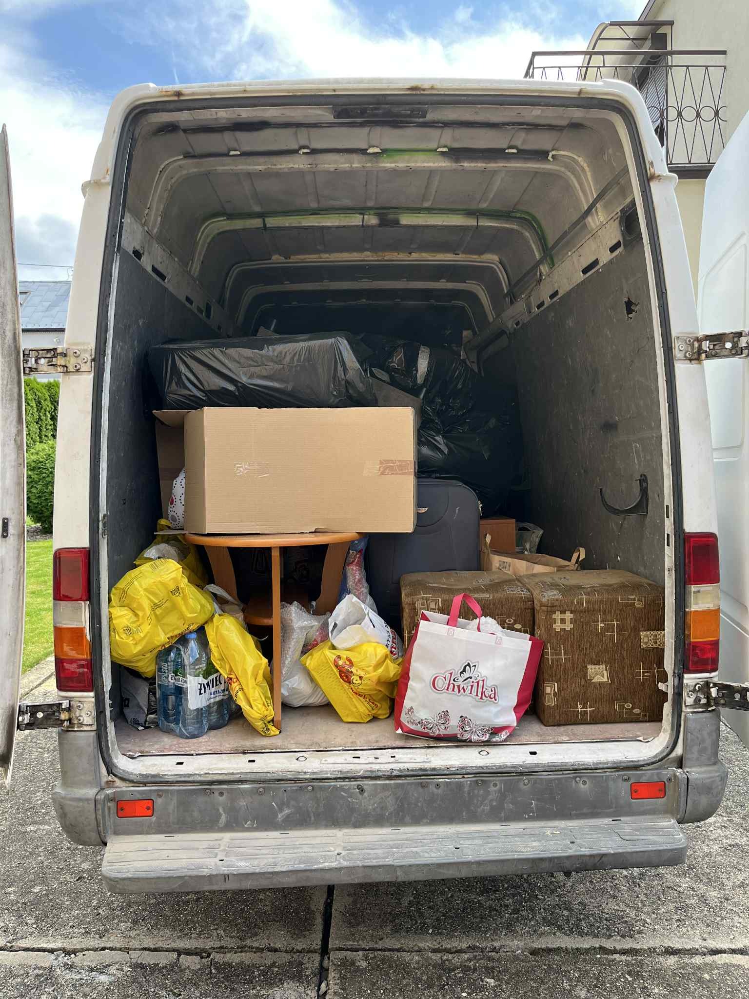 Wnętrze białego busa wypełnione różnorodnymi przedmiotami, w tym kartonami, torbami z zakupami i meblami, przygotowanymi do transportu. Widok z tyłu pojazdu.