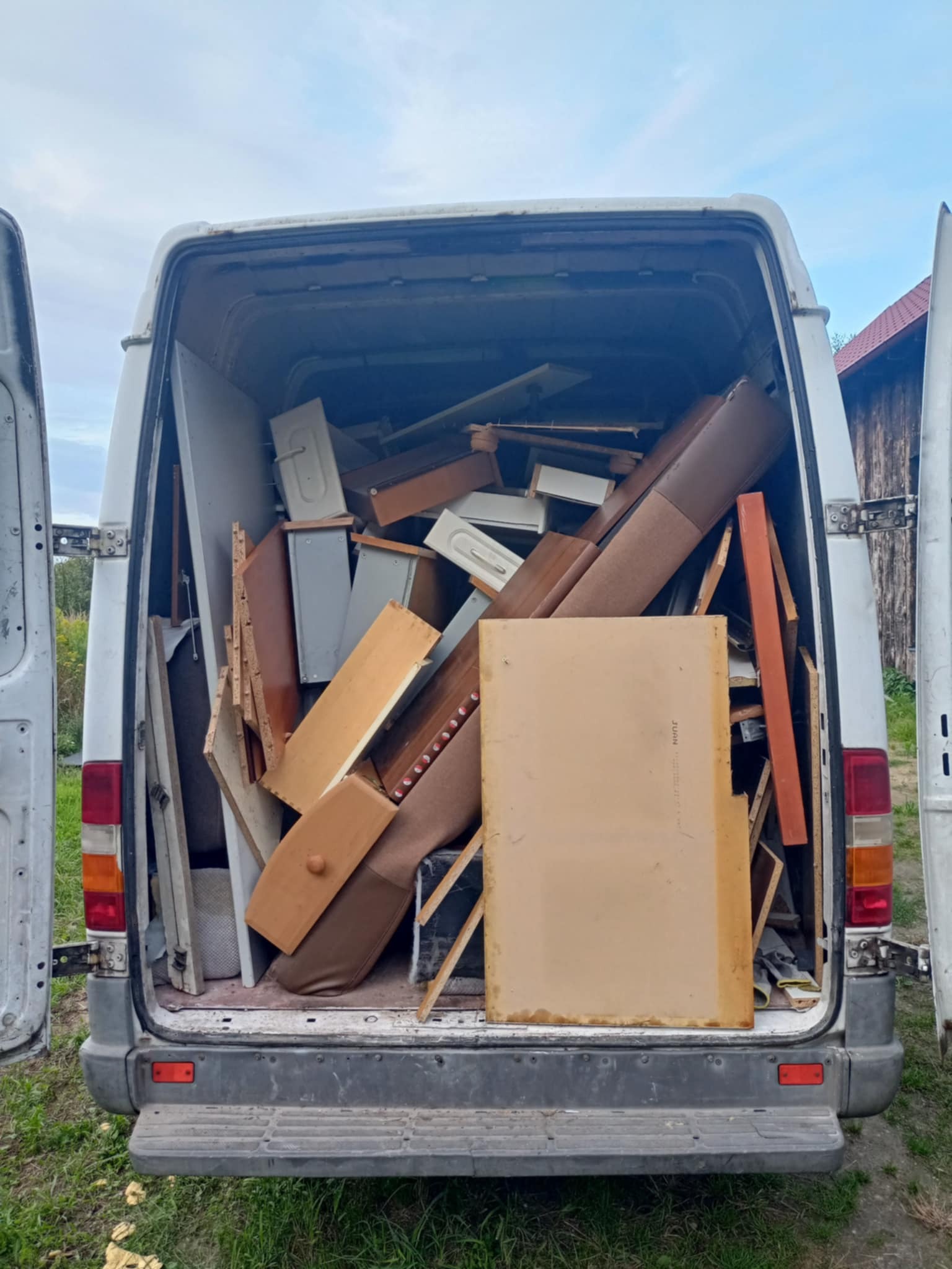 Wnętrze białego busa wypełnione meblami i elementami wyposażenia, prawdopodobnie podczas opróżniania mieszkania lub transportu. Widoczne otwarte drzwi pojazdu.