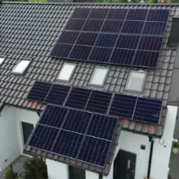 Instalacja paneli słonecznych na dachu domu jednorodzinnego z oknami dachowymi. Widok z góry, dachówka w odcieniach szarości, biała elewacja.