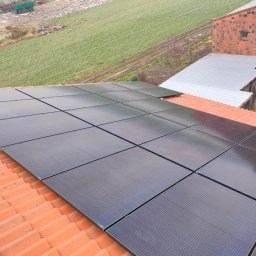 Zbigniew Kobus EXEO - Czarne panele słoneczne na pomarańczowym dachu z dachówki ceramicznej, na tle zielonego pola i budynków gospodarczych w pochmurny dzień.