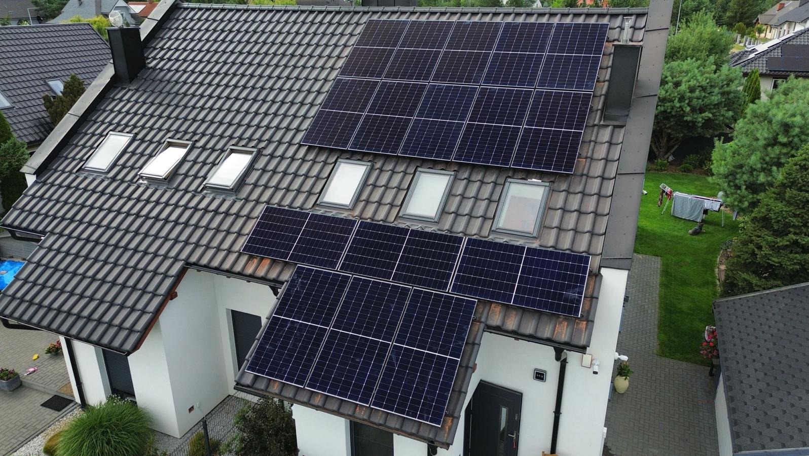 Instalacja paneli słonecznych na dachu domu jednorodzinnego z oknami dachowymi. Widok z góry, dachówka w odcieniach szarości, biała elewacja.