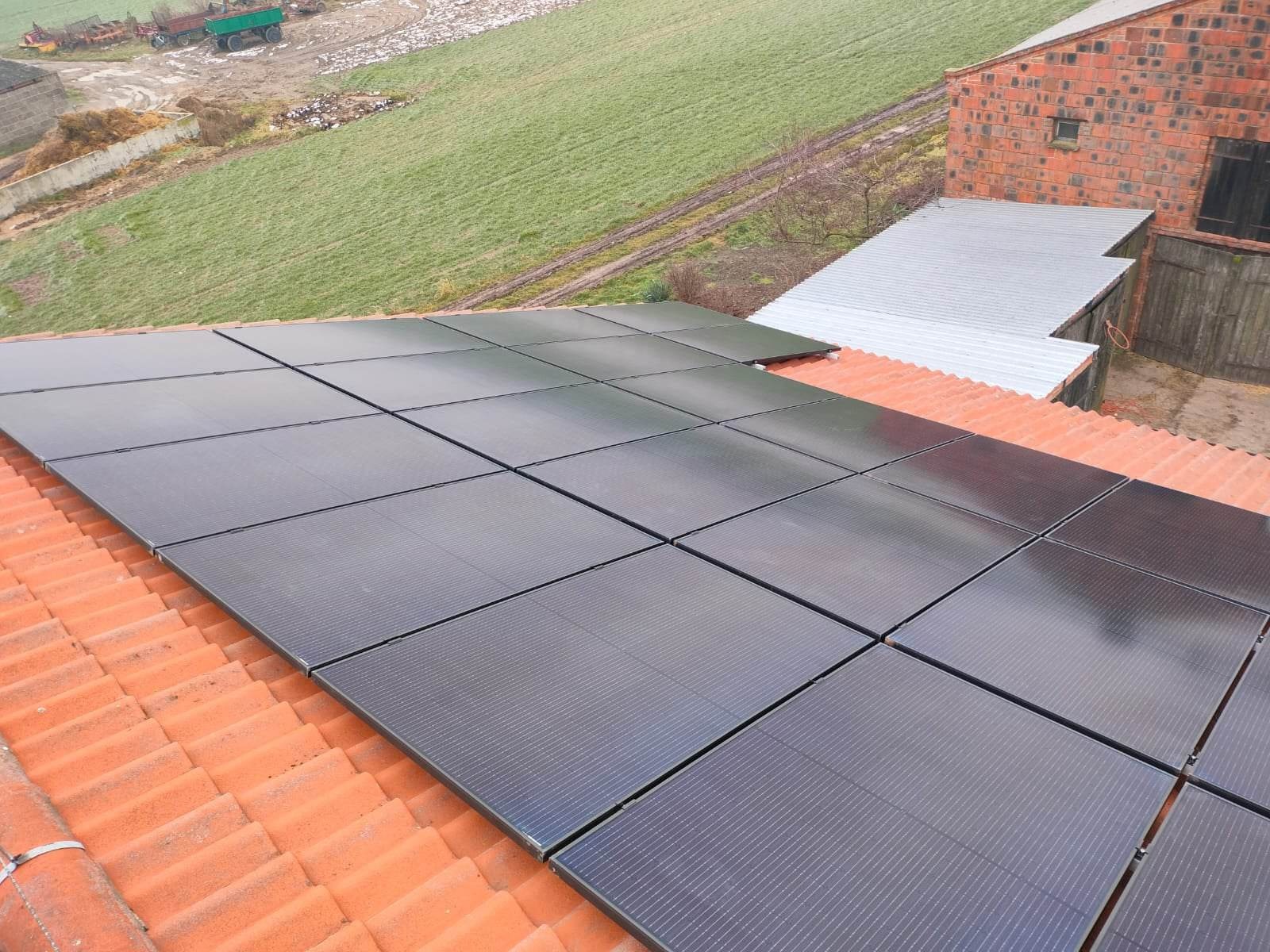 Czarne panele słoneczne na pomarańczowym dachu z dachówki ceramicznej, na tle zielonego pola i budynków gospodarczych w pochmurny dzień.