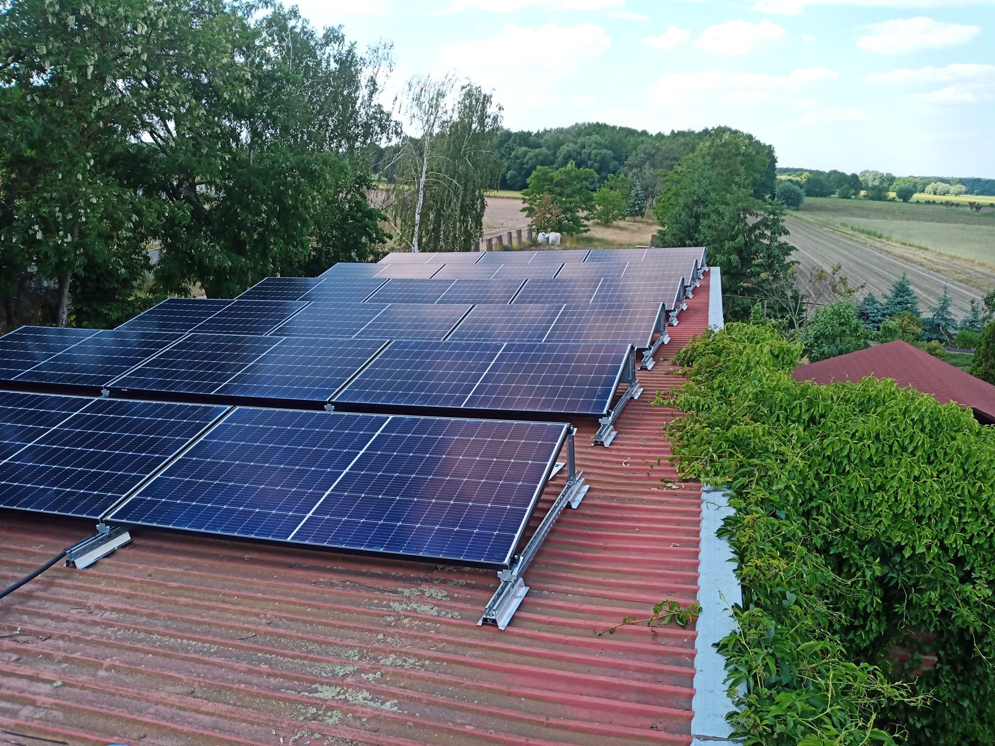 Instalacja paneli słonecznych na czerwonym dachu z blachy falistej, widok z góry, z zielenią w tle i fragmentem pola uprawnego w oddali. Moduły ułożone rzędami.