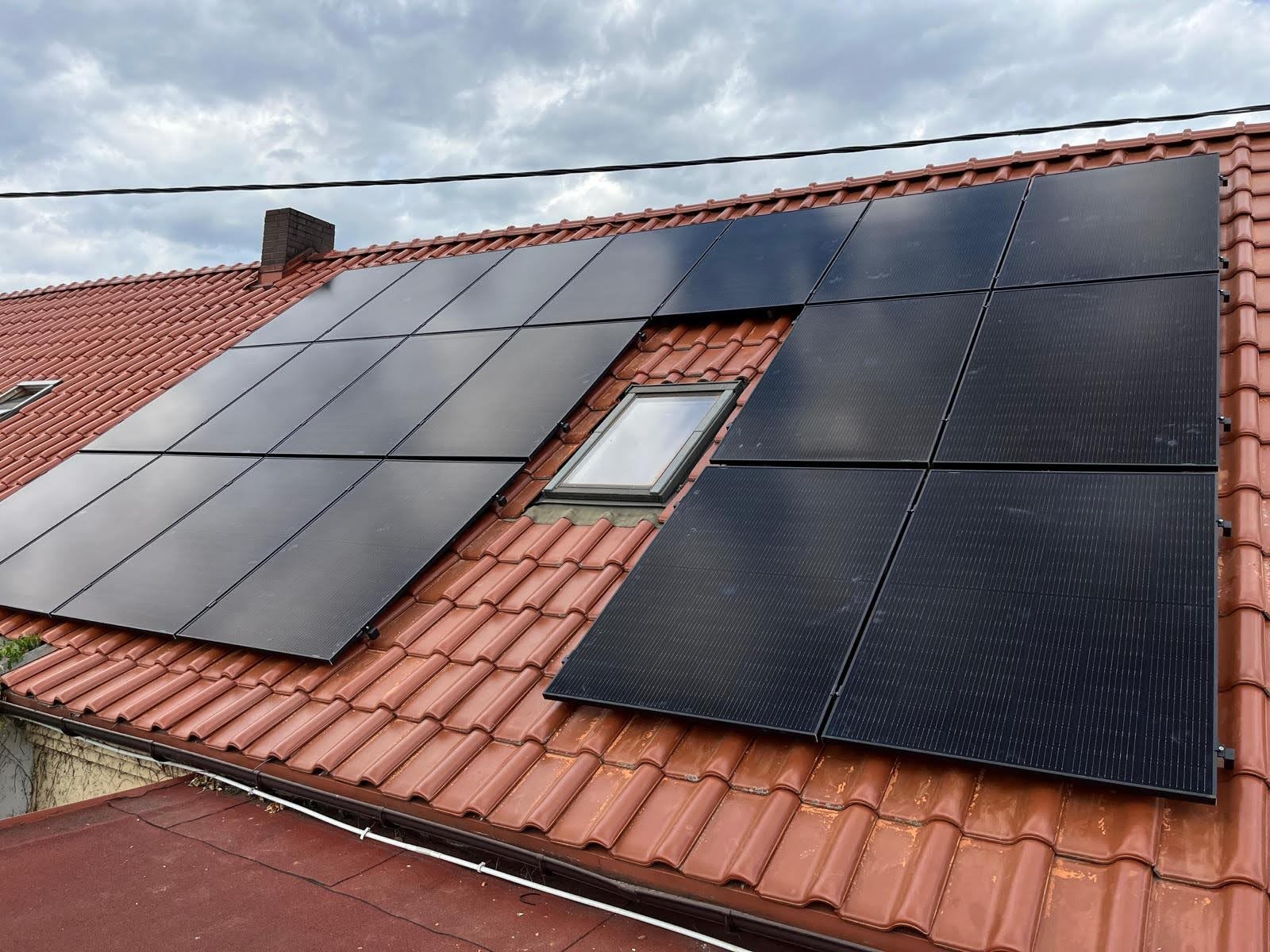 Czarne panele słoneczne na czerwonym dachu domu z oknem dachowym, pochmurne niebo w tle. Widok z dołu.