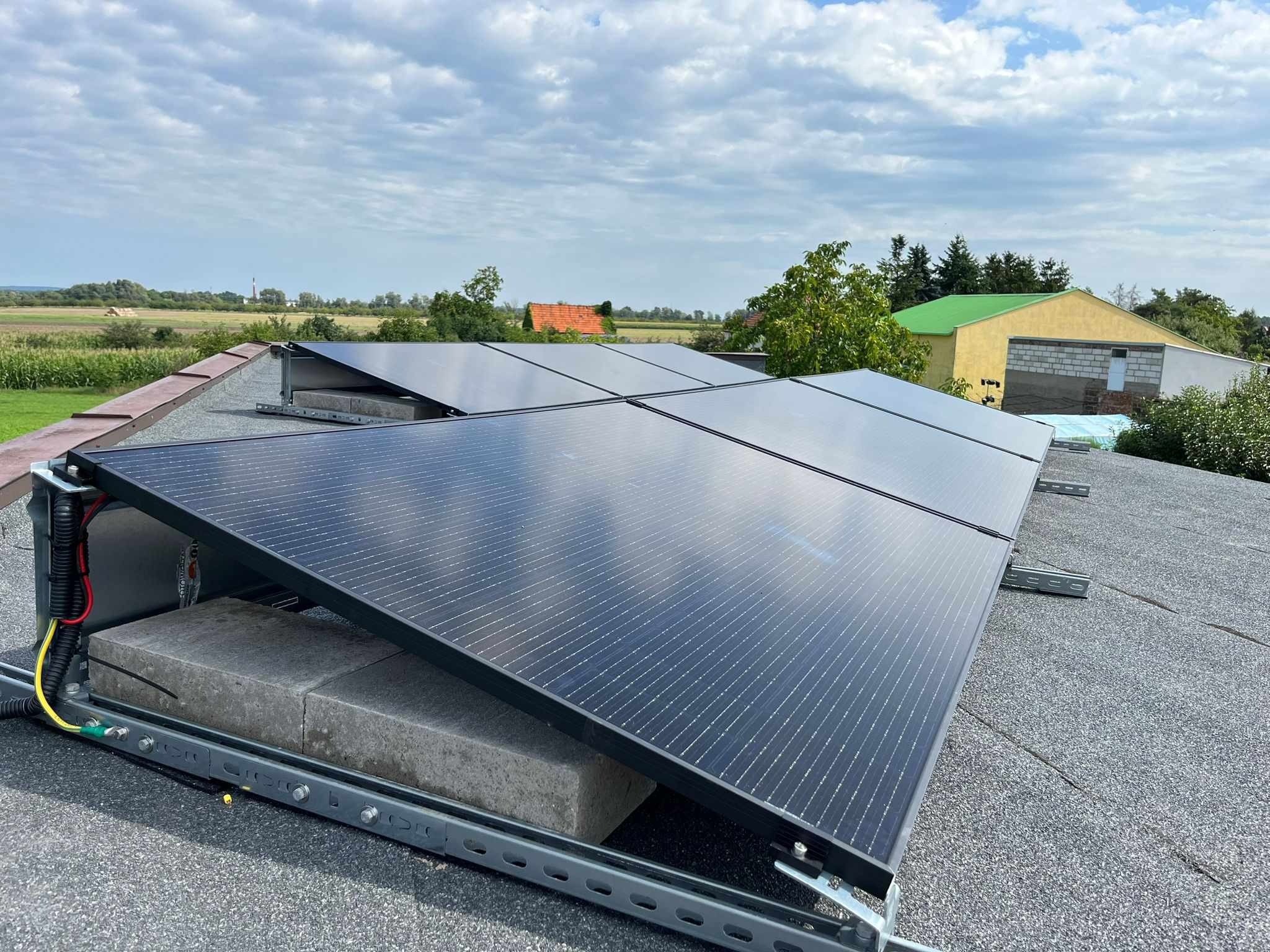Panele fotowoltaiczne na dachu pokrytym papą, zamontowane na betonowych blokach z widoczną instalacją elektryczną i fragmentem krajobrazu w tle.