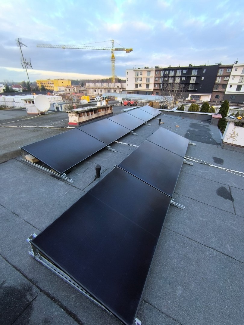 Montaż paneli słonecznych na dachu budynku z widokiem na dźwig budowlany i zabudowę miejską w tle, uchwycone w pochmurny dzień.