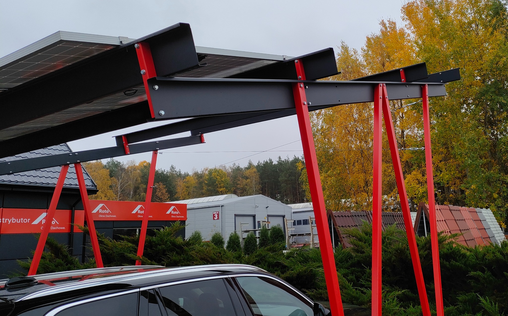 Zadaszenie fotowoltaiczne nad miejscem parkingowym, wsparte na czerwonych słupach. W tle budynki i drzewa w jesiennych barwach.