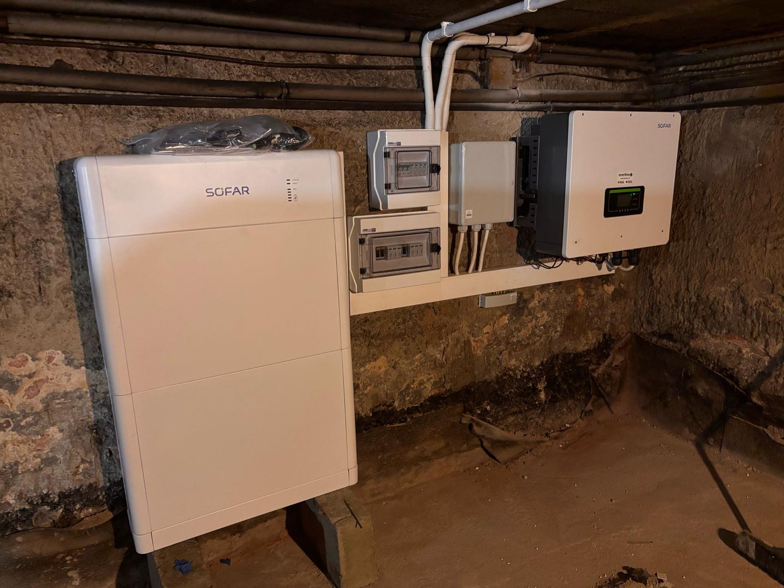 Instalacja magazynu energii Sofar w piwnicy: biała obudowa, rozdzielnice i inwerter na ścianie z rurami. Widoczne kable i betonowe podpory pod urządzeniem.