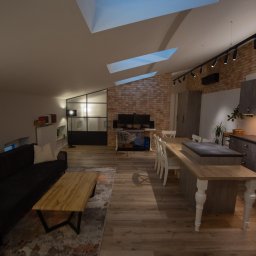 Mirosław Wesołowski - Przestronne wnętrze mieszkania z ceglaną ścianą, oświetleniem punktowym i drewnianą podłogą. Widoczny salon z sofą, stół jadalny i część kuchenna. Charakterystyczne okna dachowe.