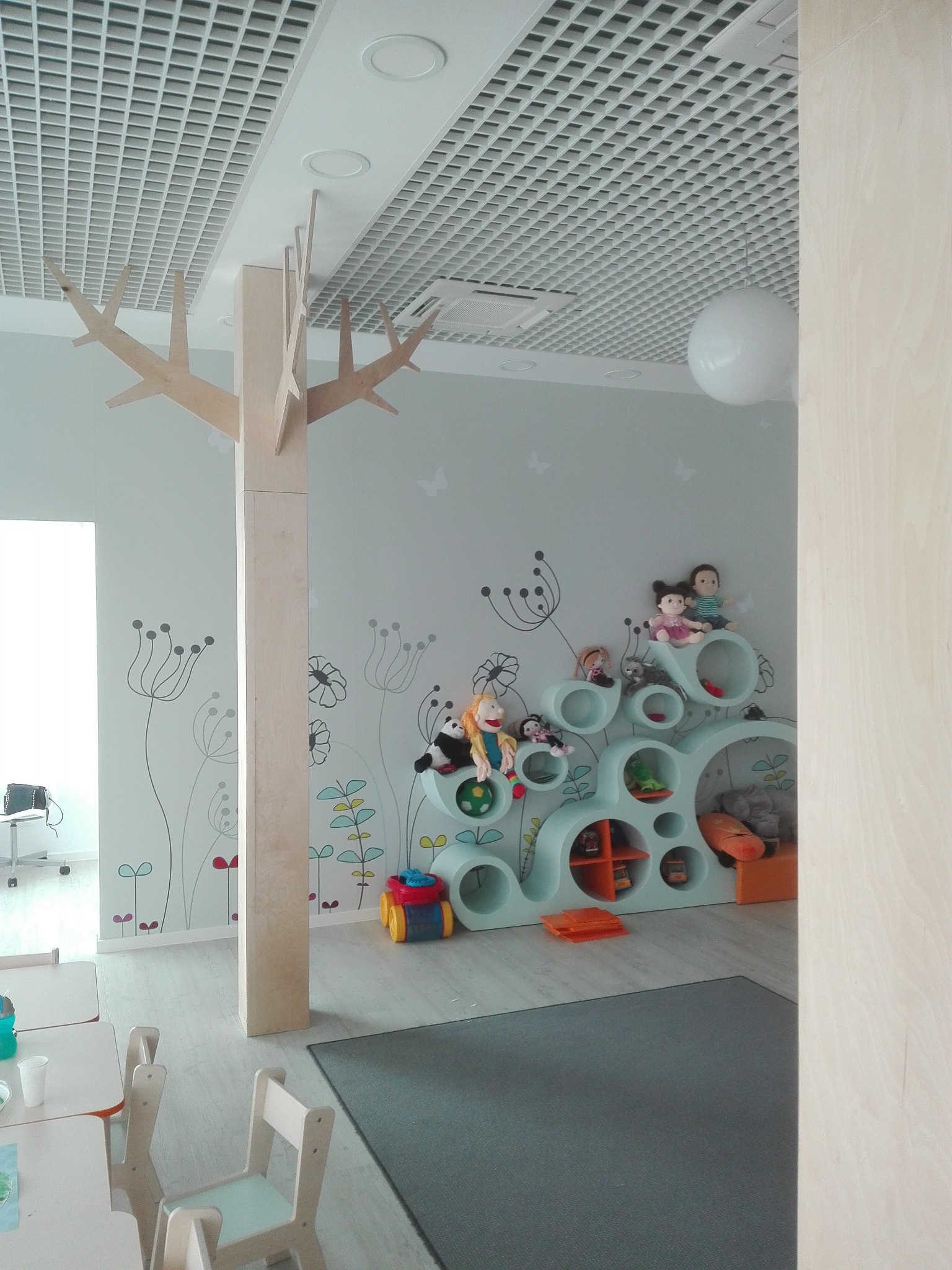 Jasne, przestronne wnętrze pokoju dziecięcego z motywem drzewa i półkami w kształcie kół. Na ścianach delikatne, florystyczne wzory i motyle. Stonowana kolorystyka.