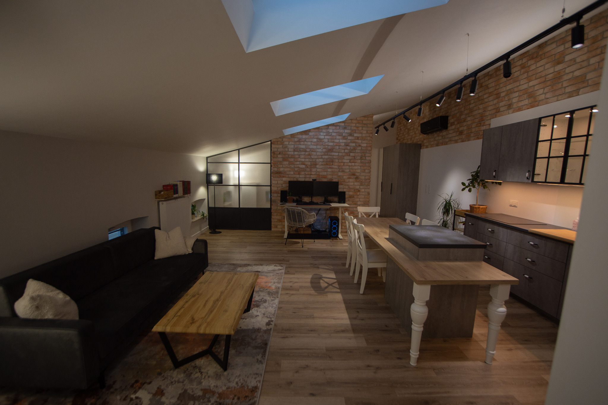 Przestronne wnętrze mieszkania z ceglaną ścianą, oświetleniem punktowym i drewnianą podłogą. Widoczny salon z sofą, stół jadalny i część kuchenna. Charakterystyczne okna dachowe.