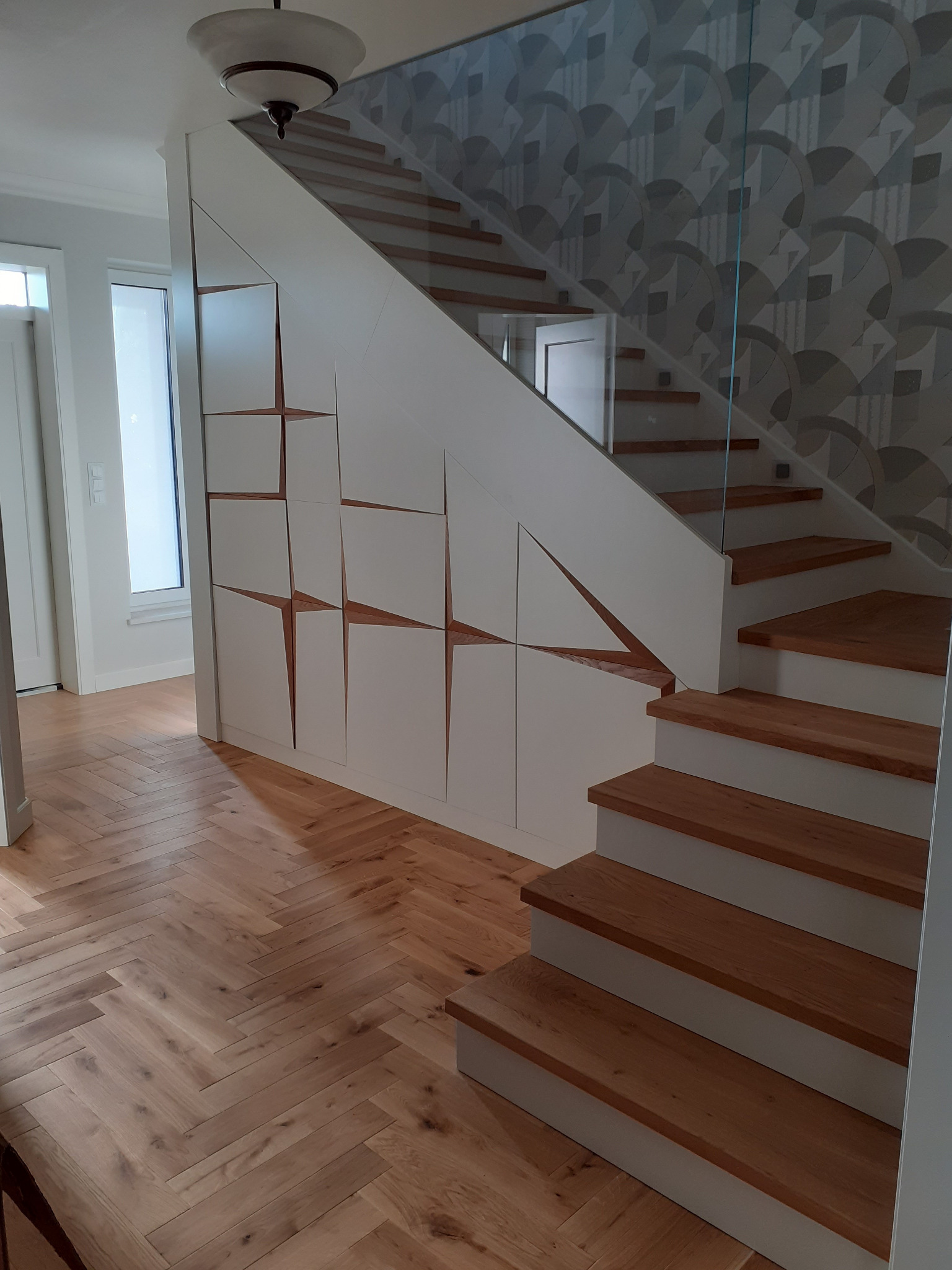 Nowoczesne schody z drewnianymi stopniami i szklaną balustradą. Pod schodami zabudowa z białymi szafkami z dekoracyjnymi, drewnianymi wstawkami. Parkiet w jodełkę na podłodze.