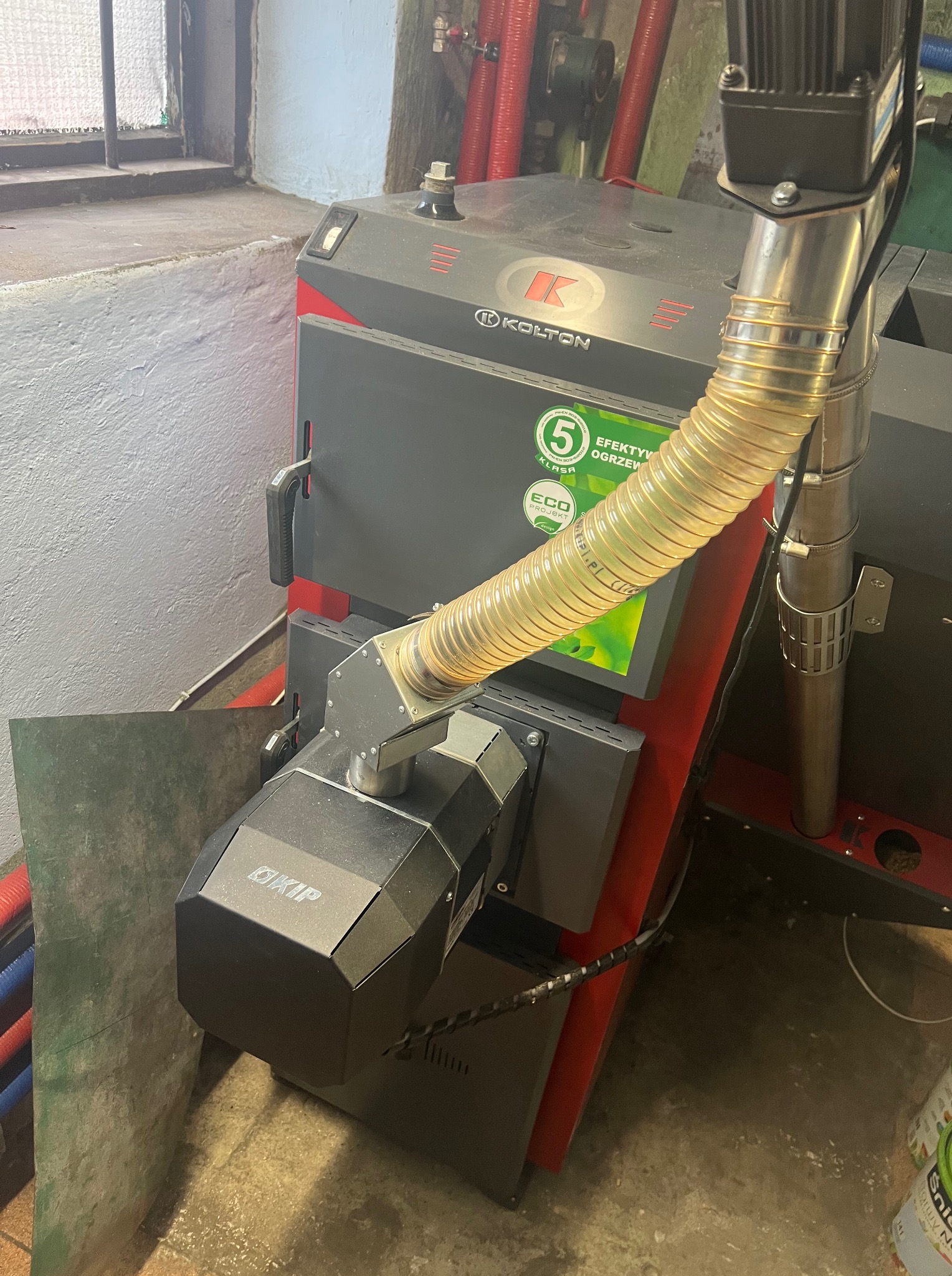 Kocioł na ekogroszek marki Kolton z podajnikiem, widoczny w kotłowni. Na kotle naklejka informująca o klasie efektywności energetycznej.