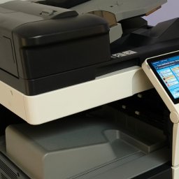 BIUROTECH - SERWIS ŁUKASZ HIPP - Wielofunkcyjna kserokopiarka biurowa z panelem dotykowym, widok z boku, szaro-biała obudowa, otwarty podajnik papieru, na tle jasnej ściany.