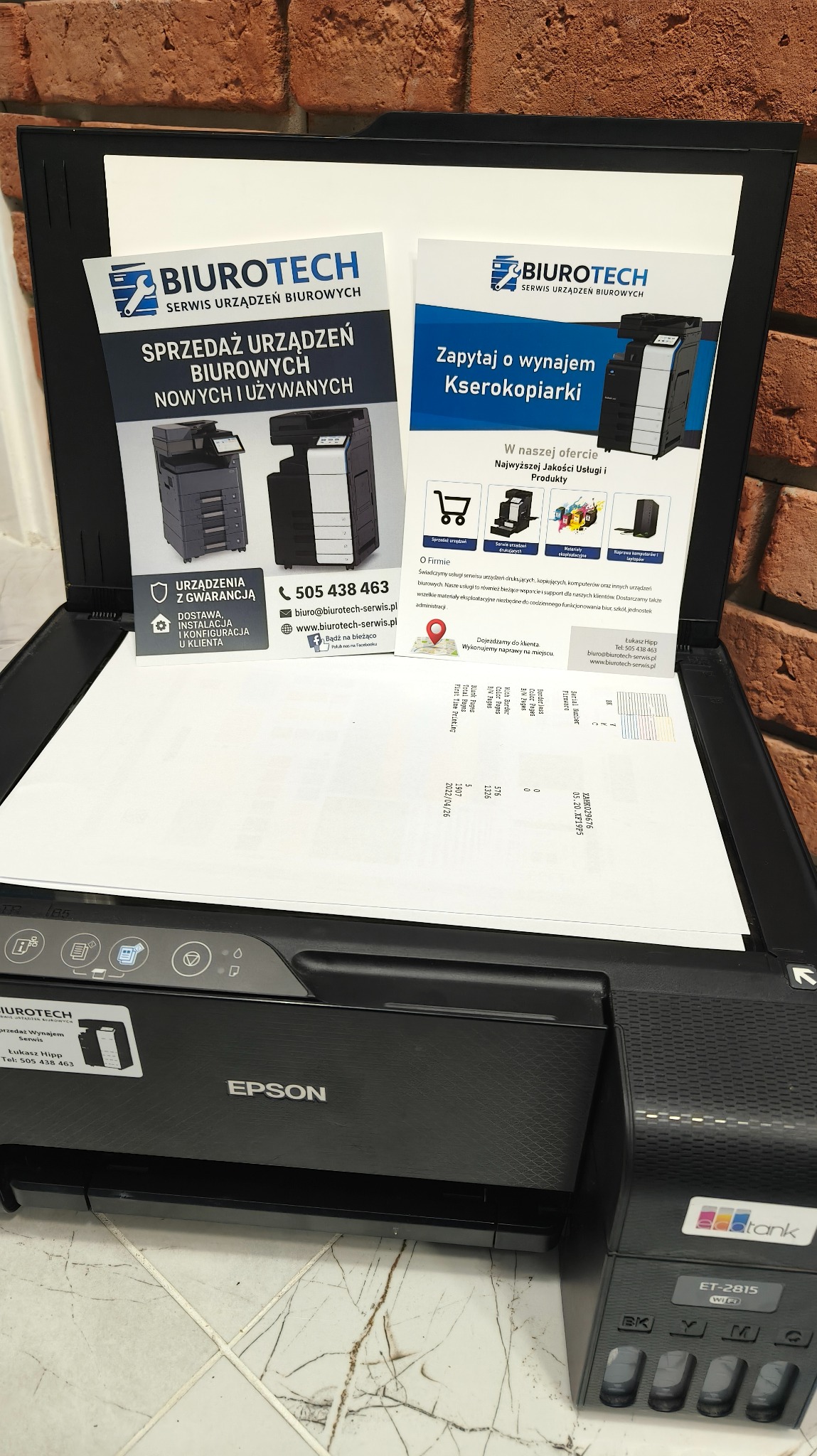 Drukarka Epson z otwartą klapą skanera, wewnątrz ulotki firmy Biurotech oferującej sprzedaż i wynajem kserokopiarek oraz serwis urządzeń biurowych. Widoczny numer telefonu.