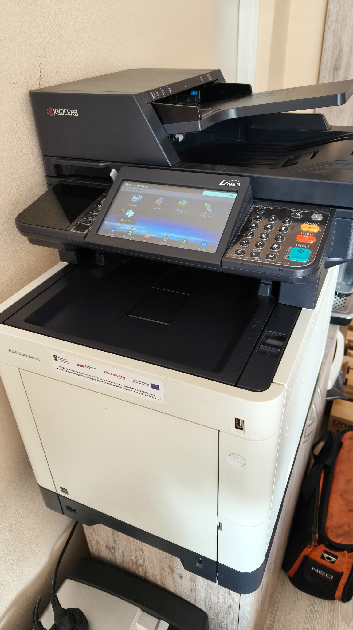 Kserokopiarka Kyocera ECOSYS M6230cidn z panelem dotykowym i przyciskami, widoczna naklejka informacyjna o dofinansowaniu z UE. Sprzęt biurowy w jasnym pomieszczeniu.