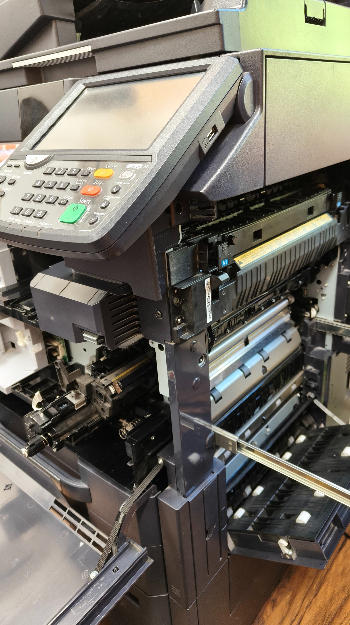 Otwarta kserokopiarka biurowa w trakcie serwisu, widoczne elementy wewnętrzne i panel sterowania z przyciskami. Sprzęt w kolorze ciemnoszarym.