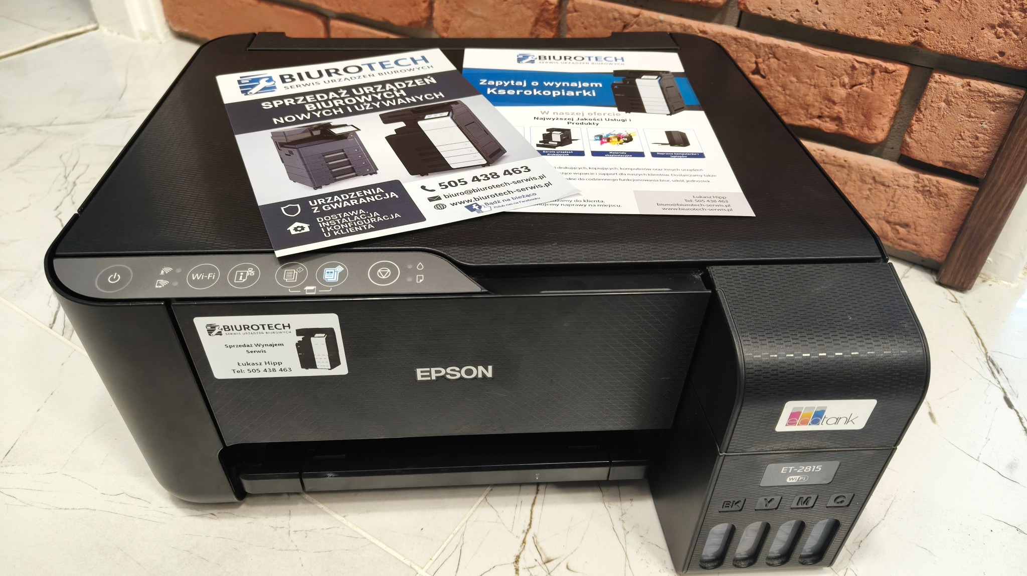 Czarna drukarka Epson ET-2815 z widocznymi pojemnikami na tusz, ulotki Biurotech z ofertą sprzedaży i wynajmu kserokopiarek, na tle ceglanej ściany.
