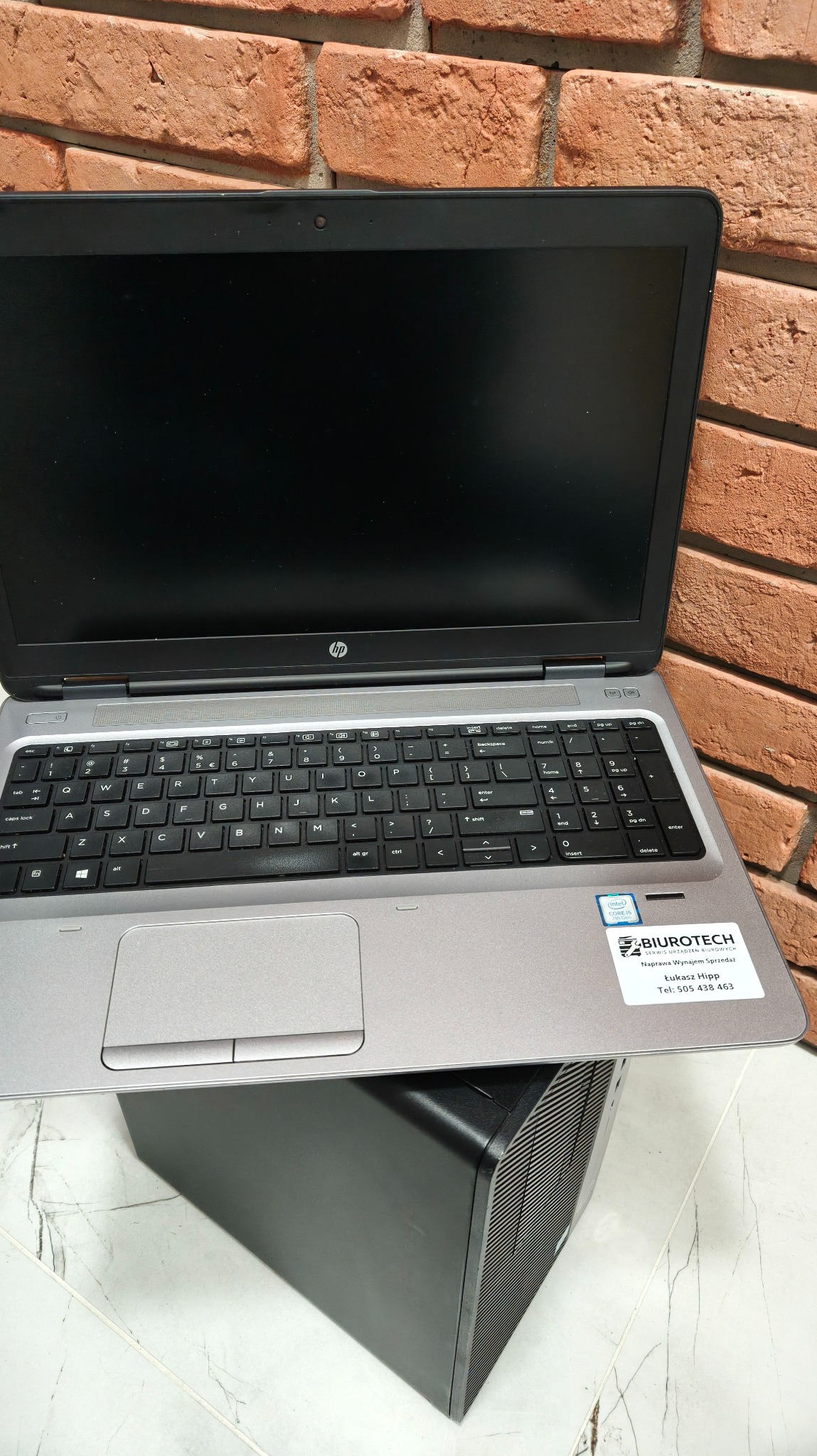 Otwarty laptop HP na obudowie komputera stacjonarnego, na tle ceglanej ściany. Naklejka serwisu Biurotech na obudowie laptopa. Widoczna klawiatura i touchpad.