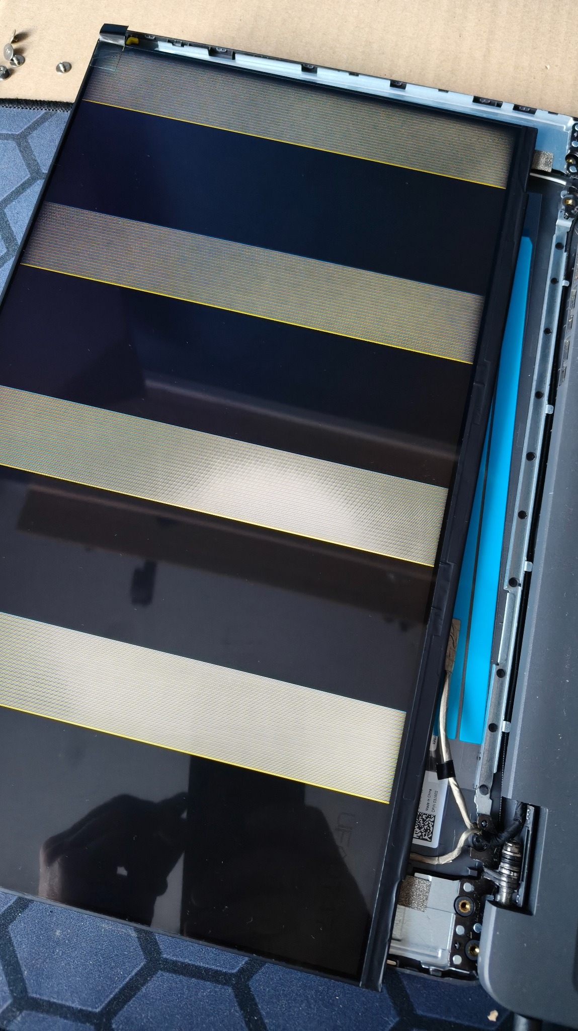 Otwarty laptop z widocznym ekranem w trakcie naprawy. Widoczne elementy elektroniki i taśmy. Specyficzny wzór na ekranie.