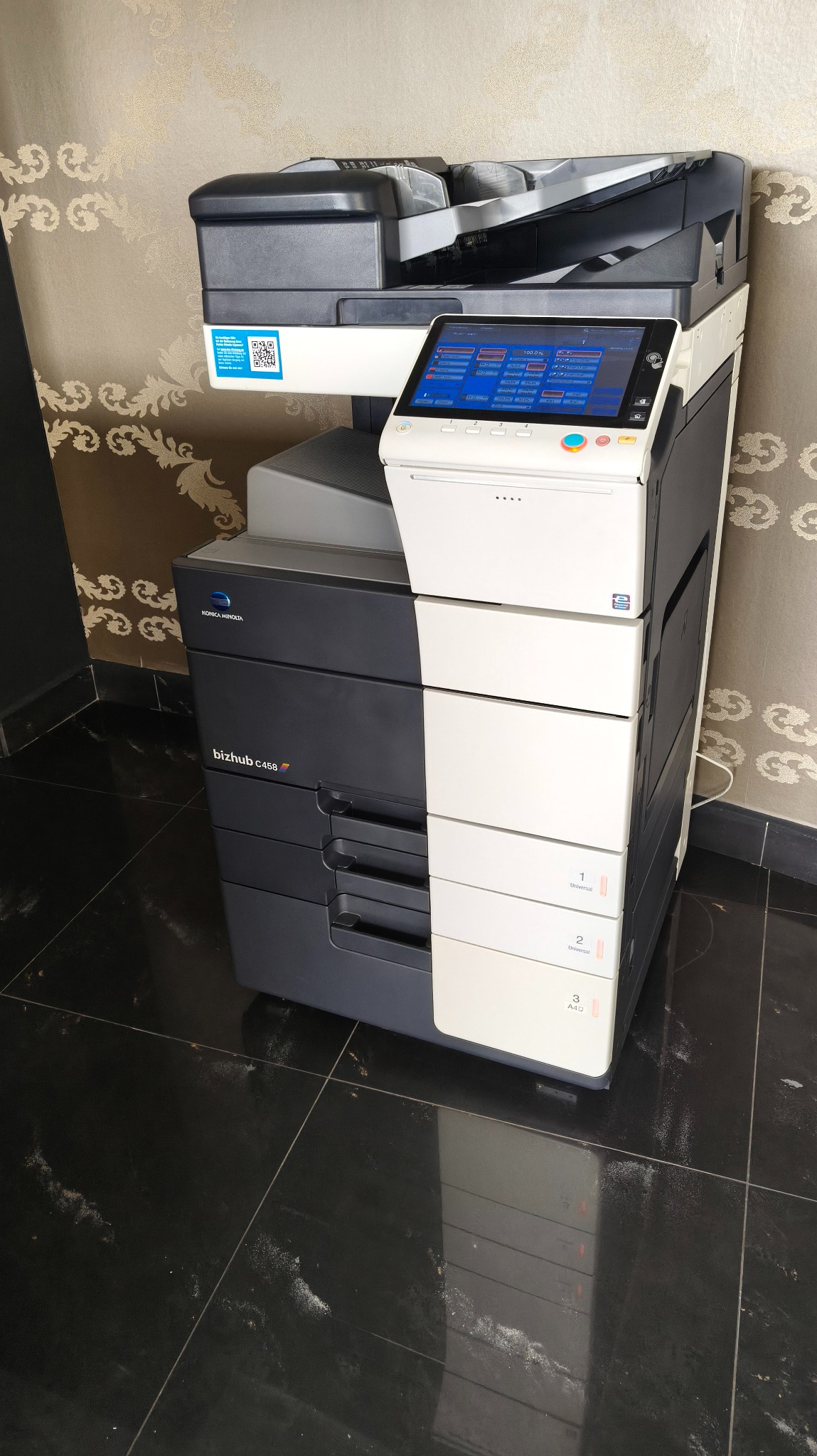 Wielofunkcyjna kserokopiarka Konica Minolta bizhub C458e, stojąca na czarnej, błyszczącej podłodze, z widocznym panelem dotykowym i podajnikami papieru.