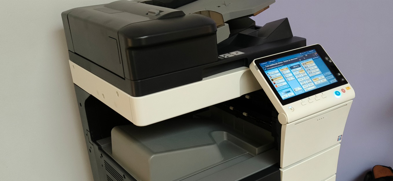 Wielofunkcyjna kserokopiarka biurowa z panelem dotykowym, widok z boku, szaro-biała obudowa, otwarty podajnik papieru, na tle jasnej ściany.