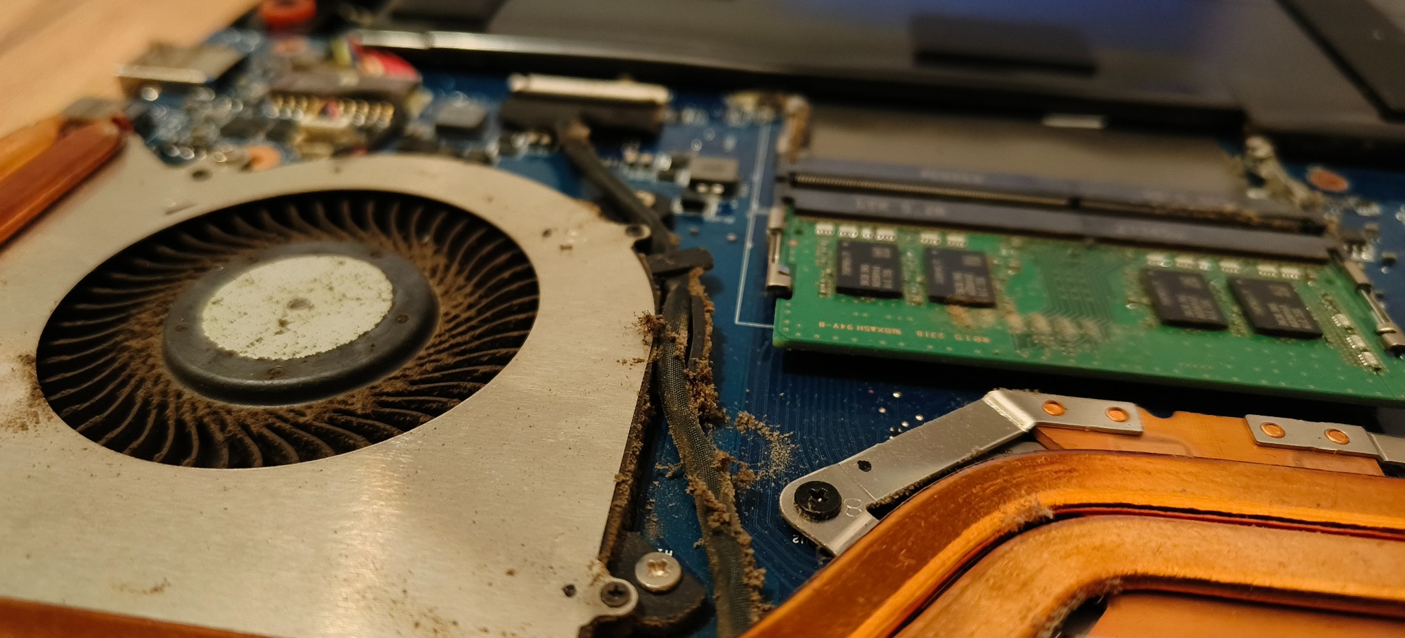 Otwarty laptop z widocznym, zakurzonym wentylatorem, pamięcią RAM i elementami płyty głównej. Detaliczne ujęcie wnętrza komputera przed czyszczeniem lub naprawą.
