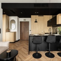 MSTUDIO KUNERT SPÓŁKA Z OGRANICZONĄ ODPOWIEDZIALNOŚCIĄ - Nowoczesne wnętrze mieszkania z aneksem kuchennym, drewnianą podłogą w jodełkę i czarnym sufitem. Widok na wyspę kuchenną i drzwi wejściowe.