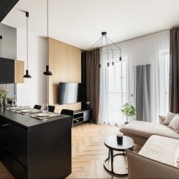 MSTUDIO KUNERT SPÓŁKA Z OGRANICZONĄ ODPOWIEDZIALNOŚCIĄ - Stylowe wykończenie wnętrza mieszkania: czarna wyspa kuchenna z blatem o fakturze kamienia, parkiet w jodełkę, beżowa sofa i nowoczesne oświetlenie.