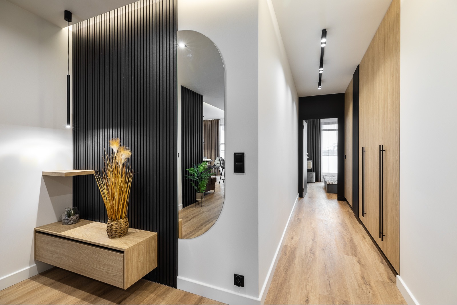 Nowoczesny przedpokój z czarną ścianą z lameli, owalnym lustrem i drewnianą komodą. W tle widoczna szafa w zabudowie i fragment sypialni. Minimalistyczny design.