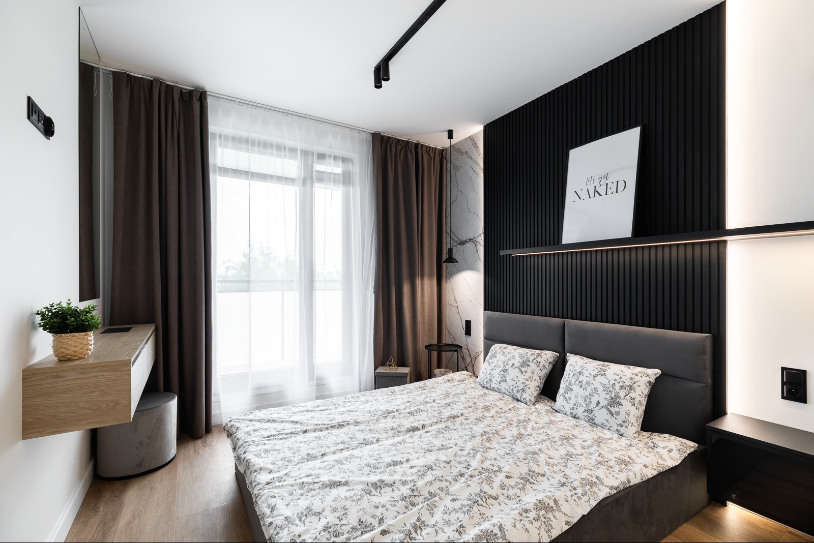 Nowoczesna sypialnia po remoncie: łóżko z szarą ramą, czarna ściana z lamelami, marmurowe akcenty, drewniana toaletka i stylowe oświetlenie.
