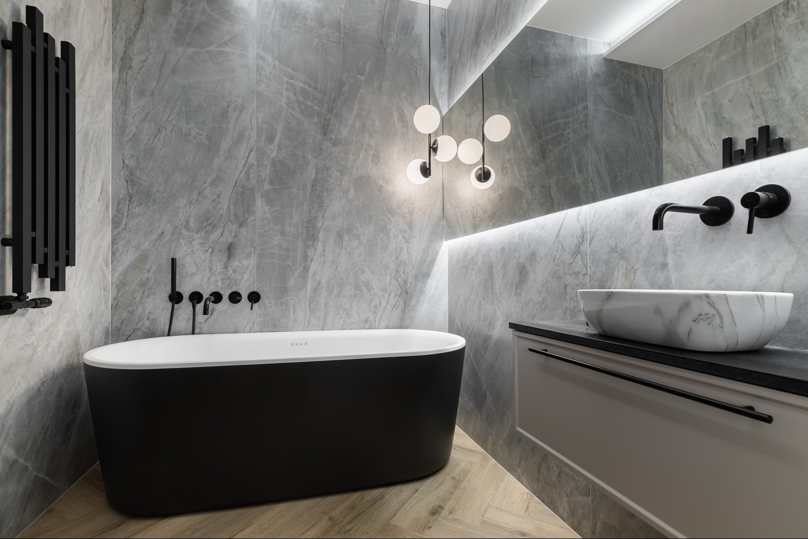 Stylowa łazienka z czarną wanną na tle marmurowych płytek, oświetlona wiszącą lampą z kulistymi kloszami i listwą LED nad lustrem. Umywalka marmurkowa.