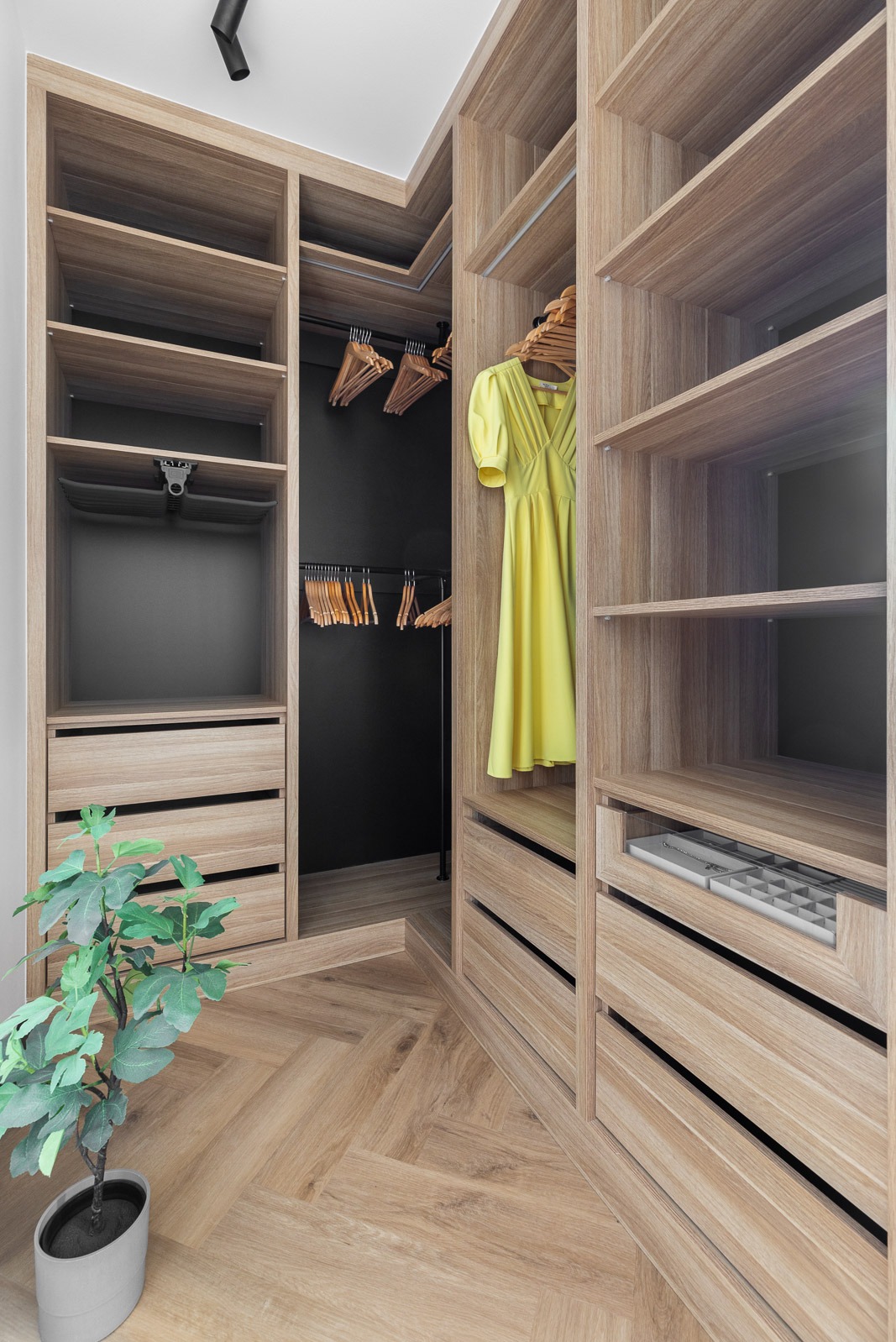 Jasna, drewniana garderoba wnękowa z czarnym tłem, żółtą sukienką i parkietem w jodełkę na podłodze, uzupełniona rośliną doniczkową.