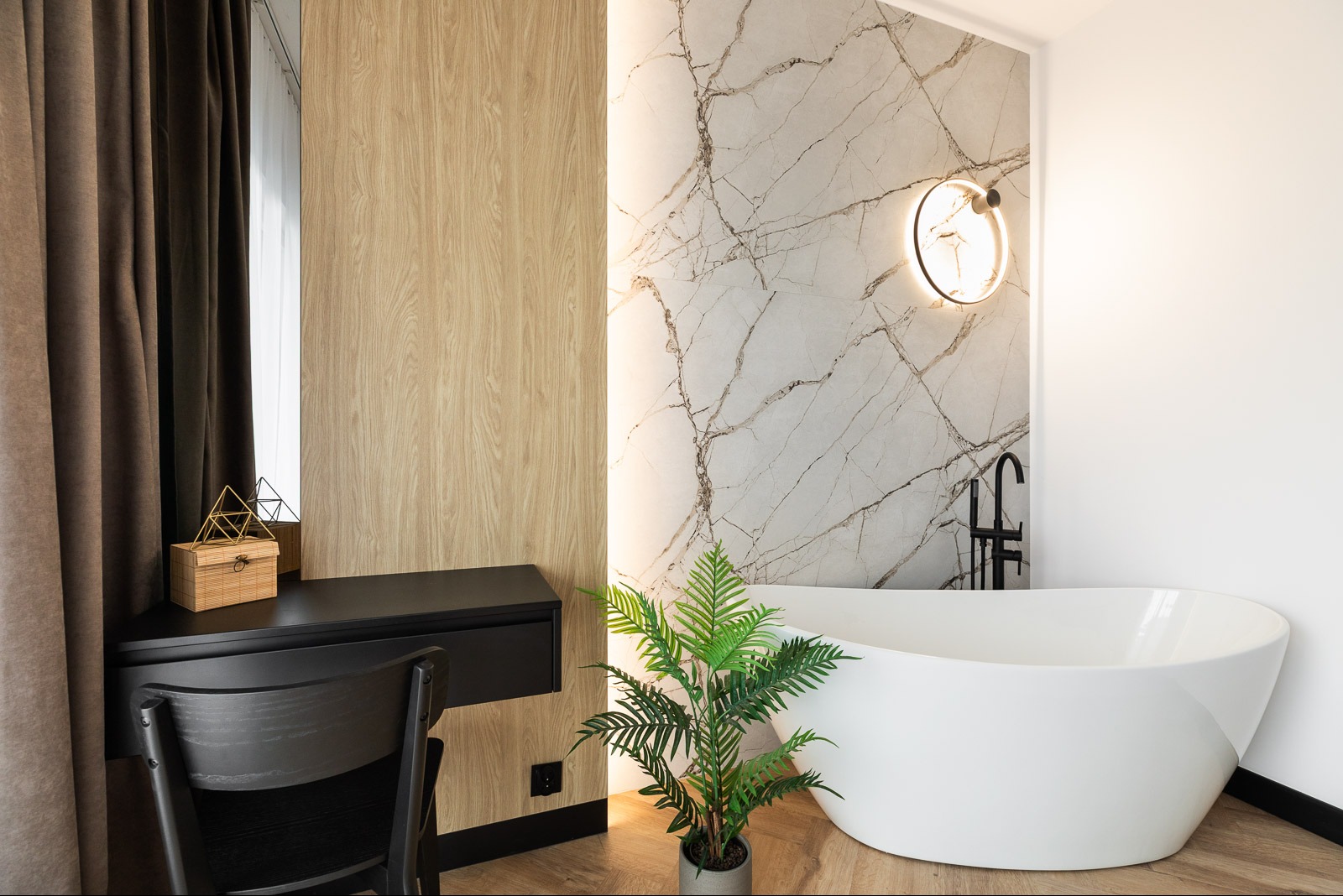 Elegancka łazienka z wolnostojącą wanną, marmurową ścianą, okrągłym lustrem z podświetleniem i drewnianą boazerią. Czarny blat z dekoracjami i zielona roślina.