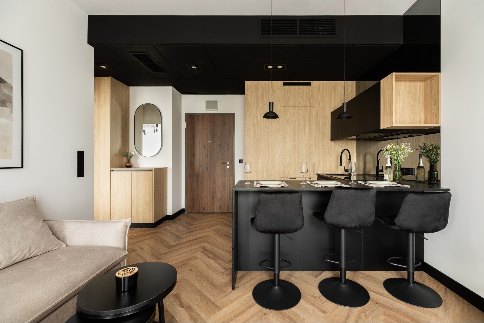 Nowoczesne wnętrze mieszkania z aneksem kuchennym, drewnianą podłogą w jodełkę i czarnym sufitem. Widok na wyspę kuchenną i drzwi wejściowe.