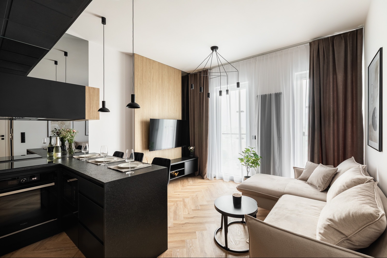 Stylowe wykończenie wnętrza mieszkania: czarna wyspa kuchenna z blatem o fakturze kamienia, parkiet w jodełkę, beżowa sofa i nowoczesne oświetlenie.