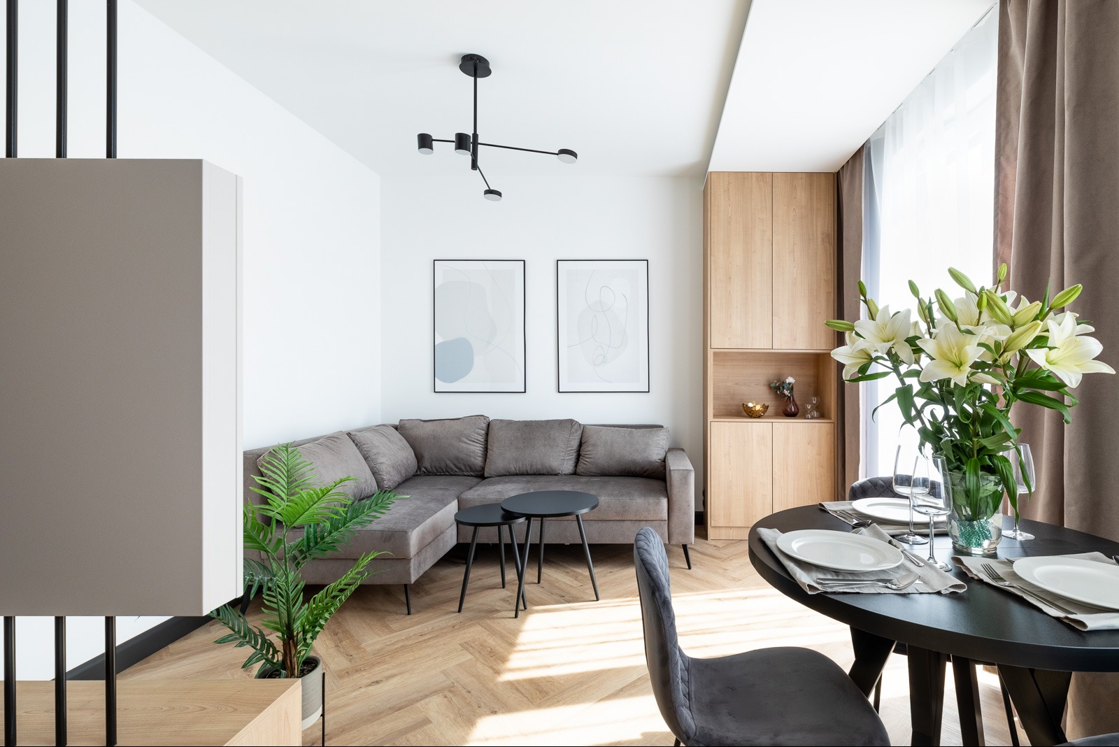 Stylowe wykończenie wnętrza salonu z narożnikiem, parkietem w jodełkę, drewnianą zabudową i czarnym stołem. Dwa abstrakcyjne obrazy na ścianie.