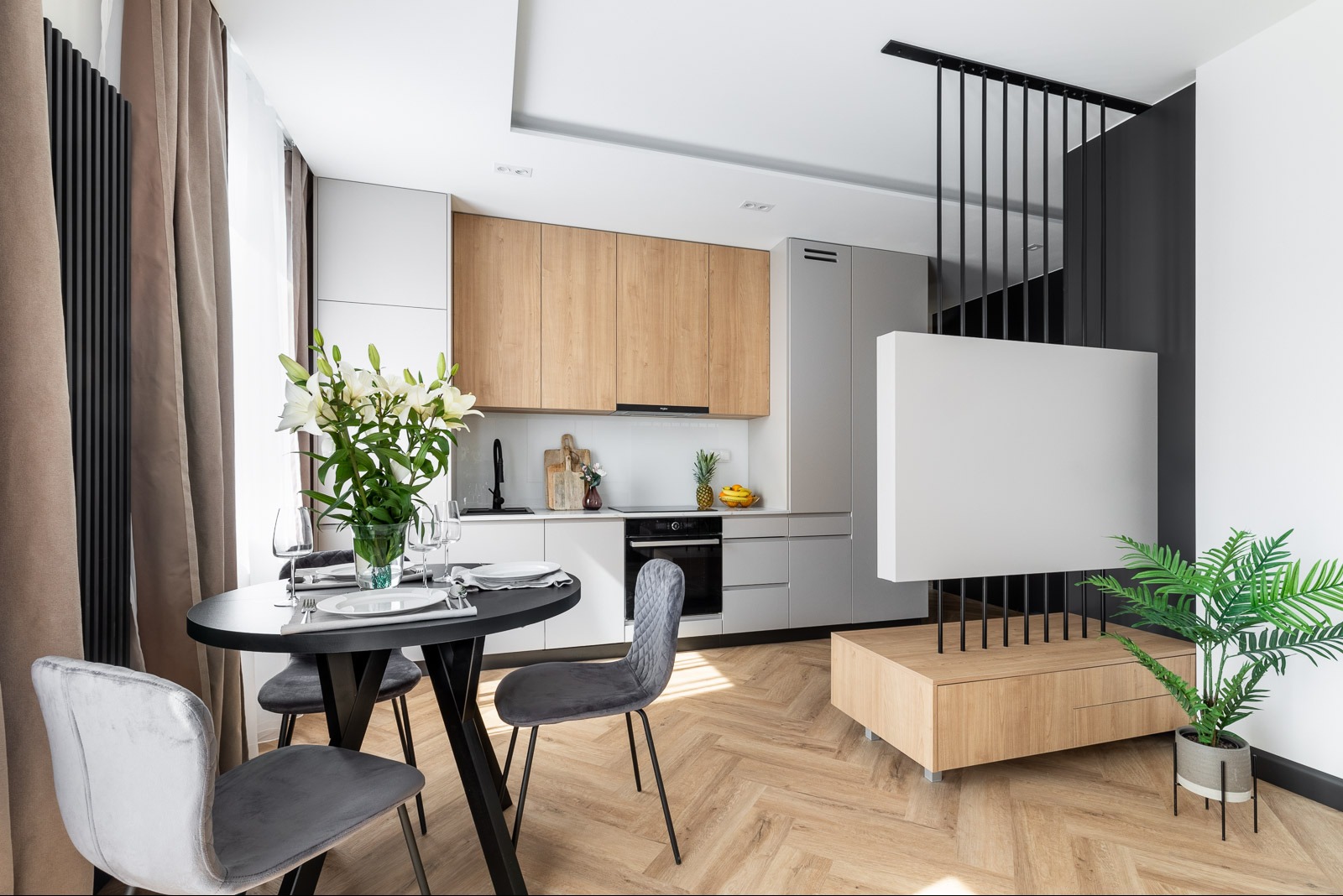 Nowoczesna kuchnia z drewnianymi i szarymi szafkami, okrągłym stołem z kwiatami oraz podłogą w jodełkę, oświetlona naturalnym światłem.