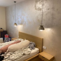 Arsen Prokopovycz - Stylowa sypialnia z łóżkiem z drewnianym zagłówkiem i lampami wiszącymi. Ściana z dekoracyjnym wykończeniem imitującym marmur. Widoczne gniazdko elektryczne.