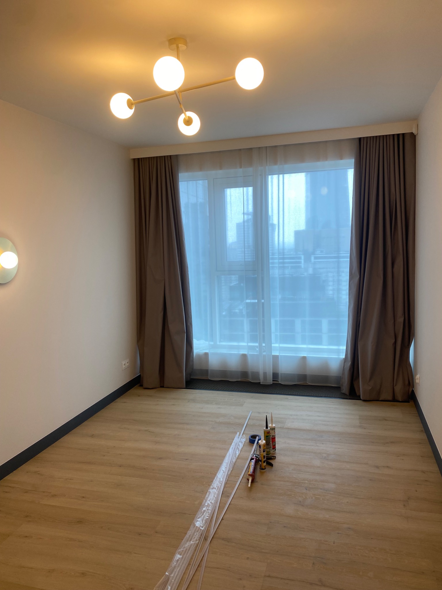 Pokój po remoncie z drewnianą podłogą, zasłonami i nowoczesną lampą. Na podłodze materiały budowlane, silikony i listwy przypodłogowe przygotowane do montażu.
