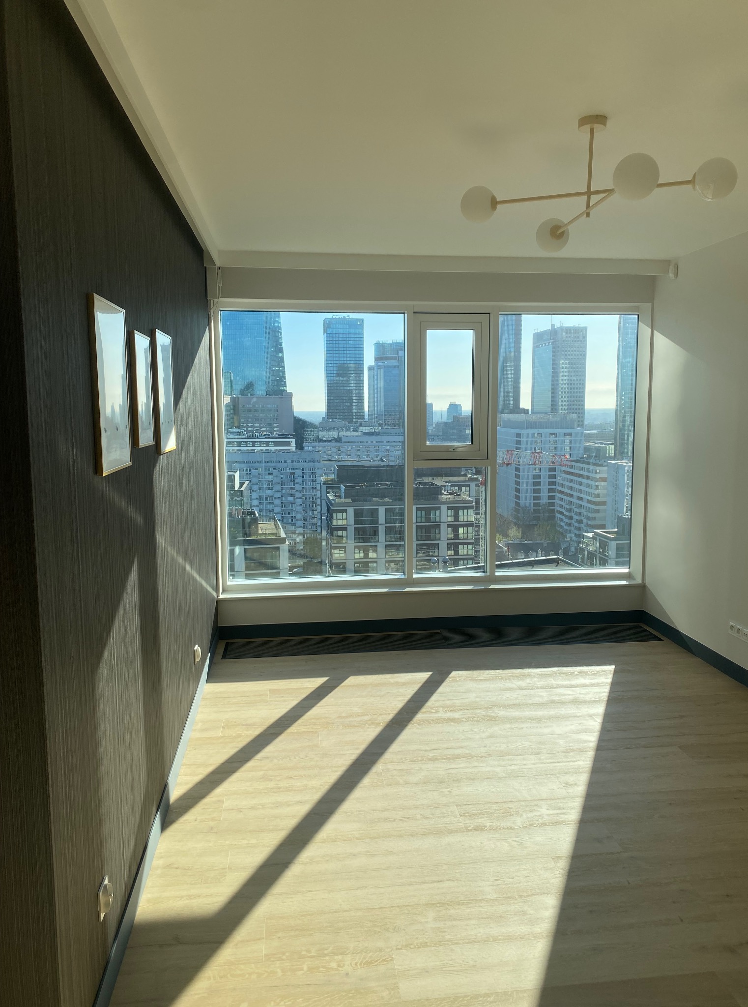 Jasne wnętrze mieszkania z parkietem i widokiem na panoramę miasta przez duże okno. Na ścianie trzy obrazy w złotych ramkach, nowoczesna lampa sufitowa.