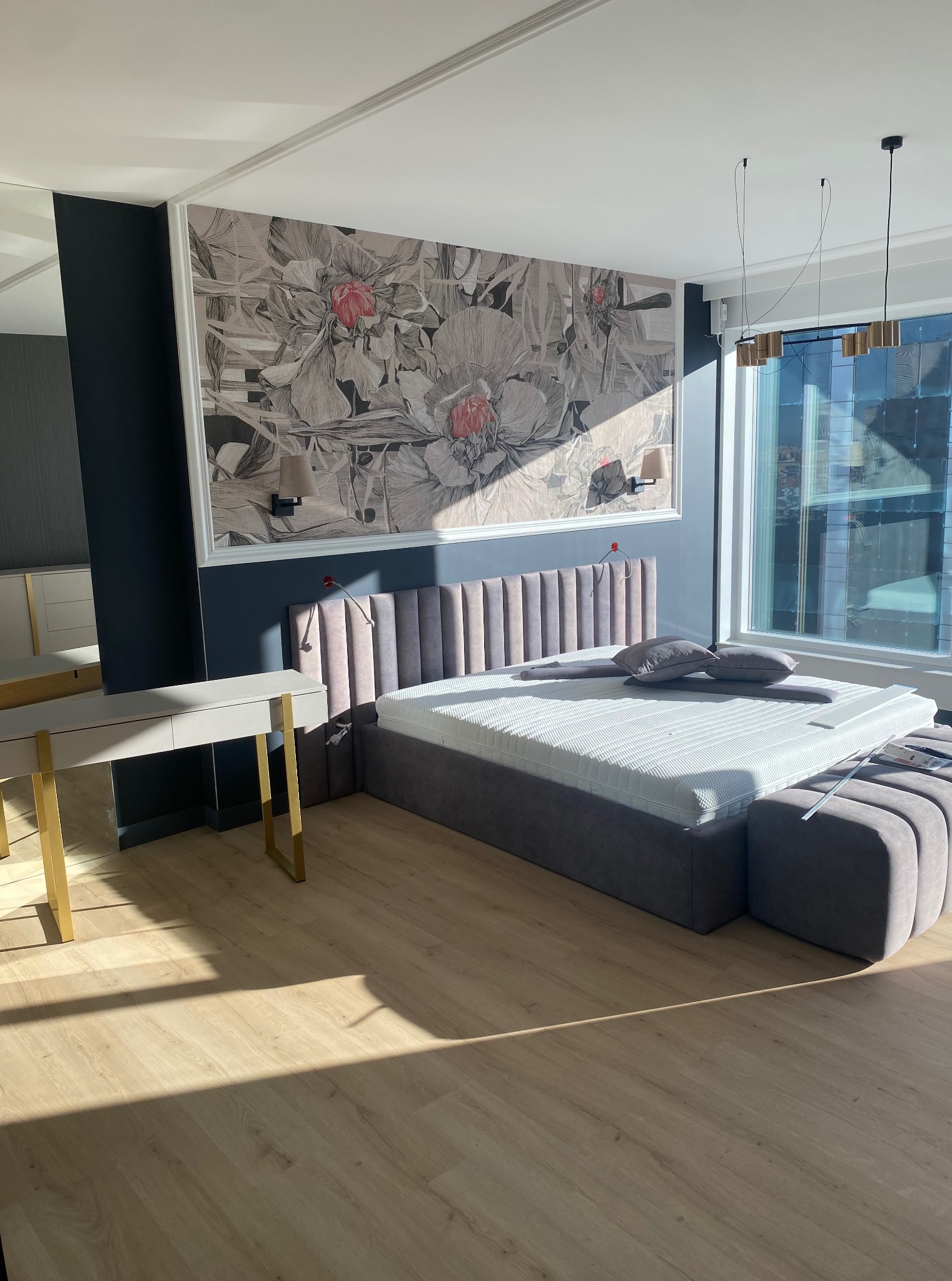 Sypialnia w nowoczesnym stylu z łóżkiem z tapicerowanym wezgłowiem, wzorzystą tapetą w kwiaty i biurkiem z metalowymi nogami, oświetlona promieniami słońca.