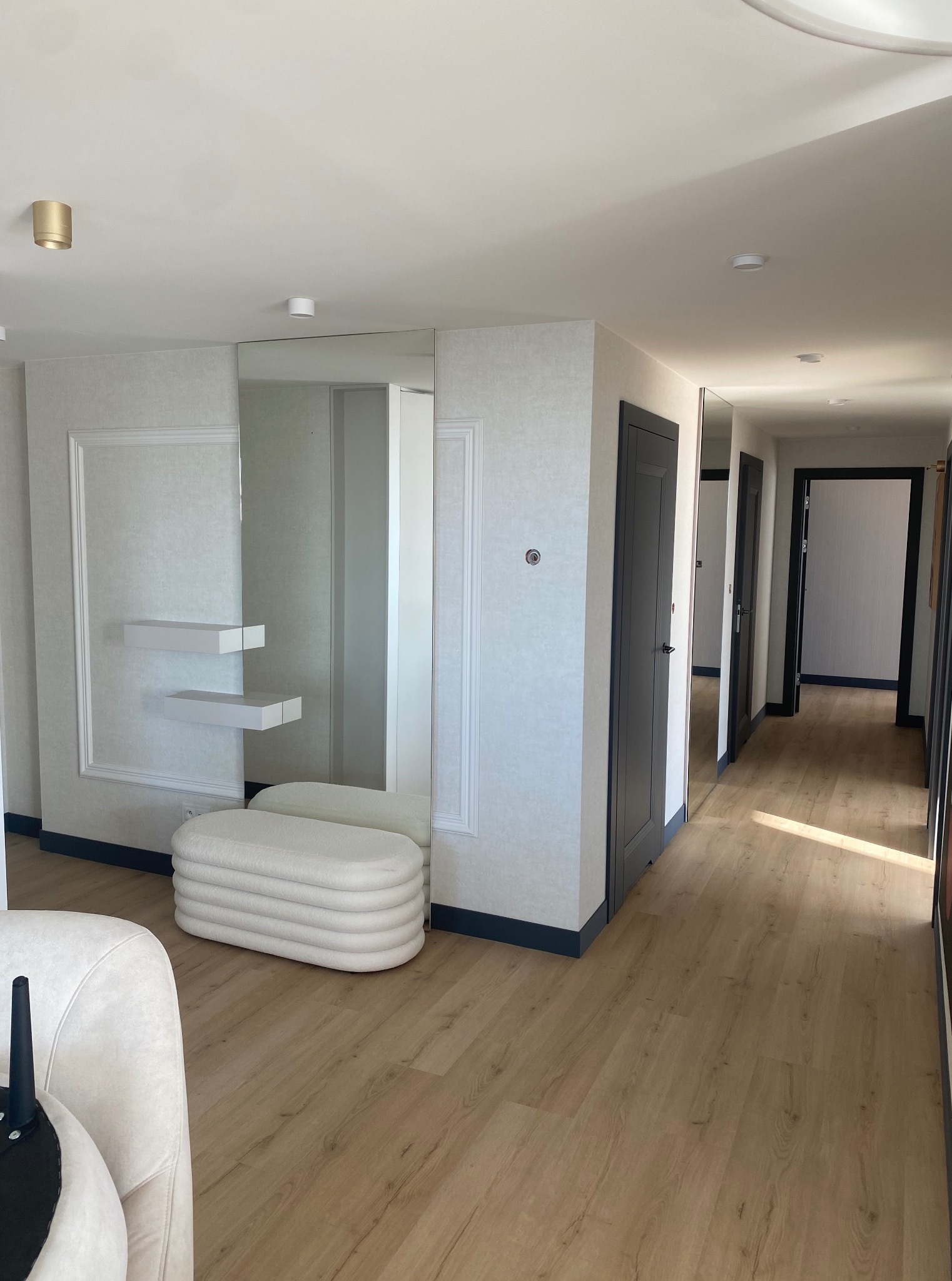 Jasne wnętrze z lustrem, półkami i pufą. Parkiet na podłodze, ciemne ramy drzwi. Dekoracyjne listwy na ścianach i minimalistyczne oświetlenie sufitowe.