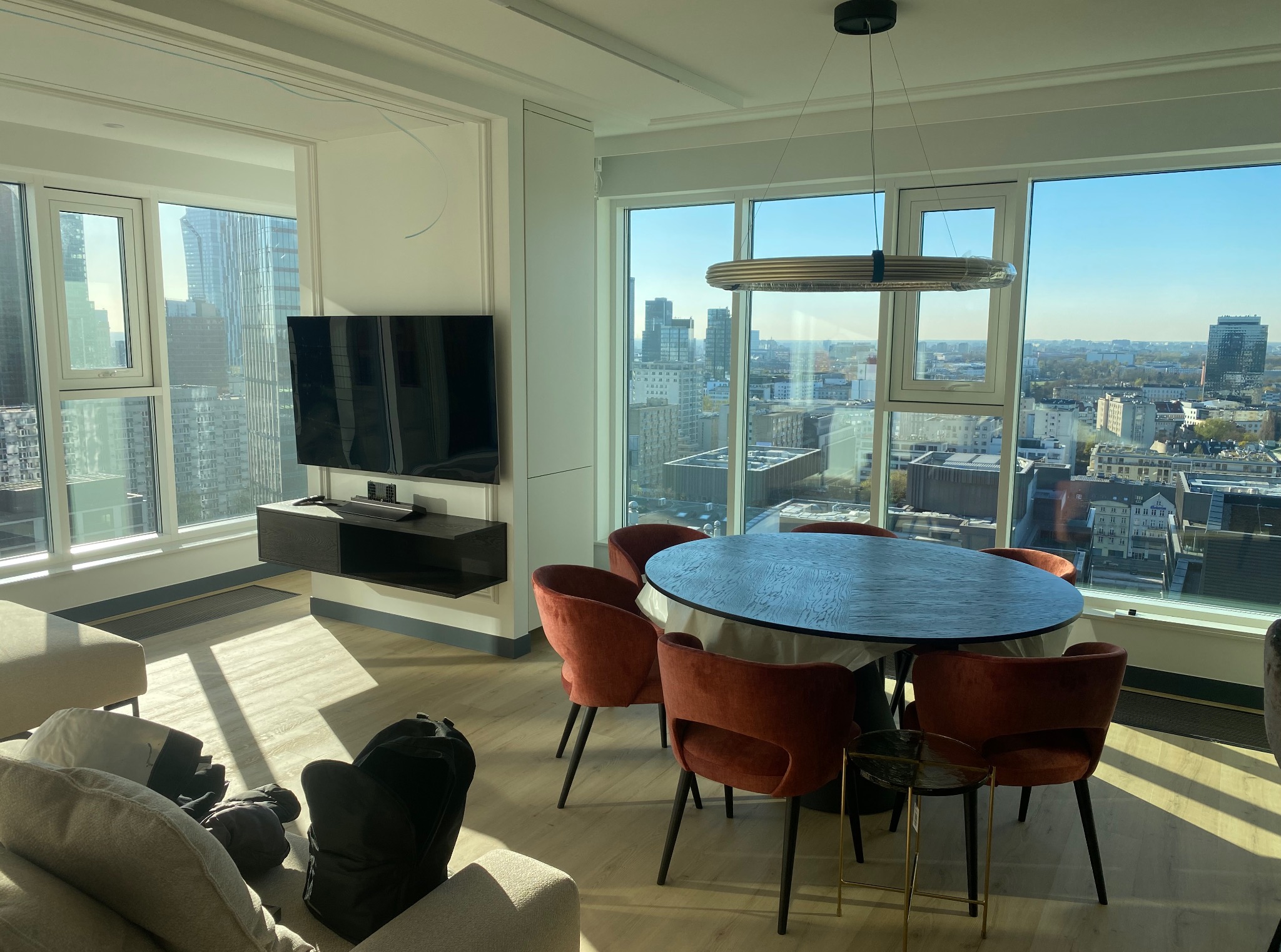 Jasne wnętrze apartamentu z widokiem na miasto. Okrągły stół z krzesłami w odcieniach czerwieni, nowoczesna lampa. Minimalistyczny wystrój, duże okna.