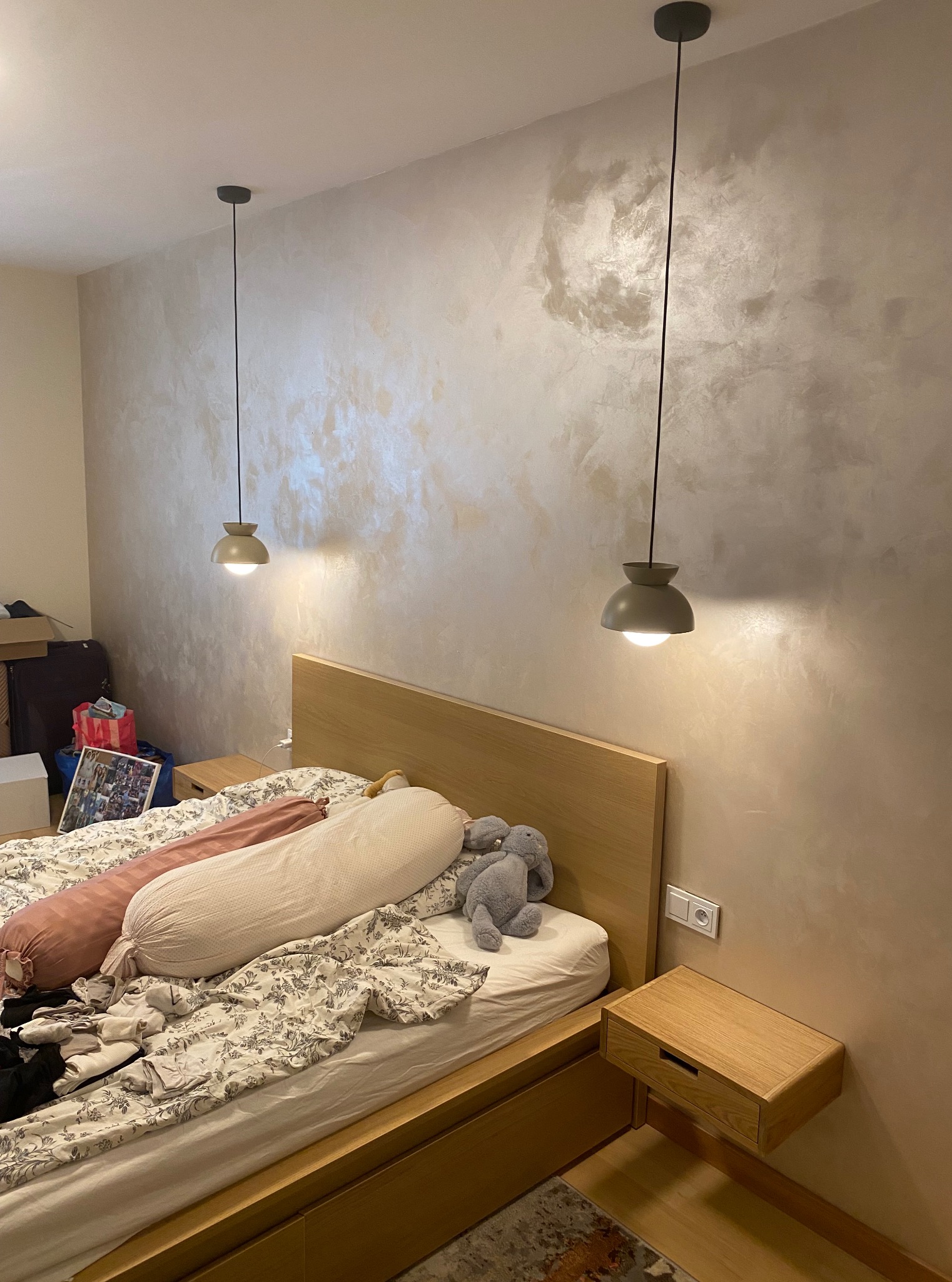 Stylowa sypialnia z łóżkiem z drewnianym zagłówkiem i lampami wiszącymi. Ściana z dekoracyjnym wykończeniem imitującym marmur. Widoczne gniazdko elektryczne.