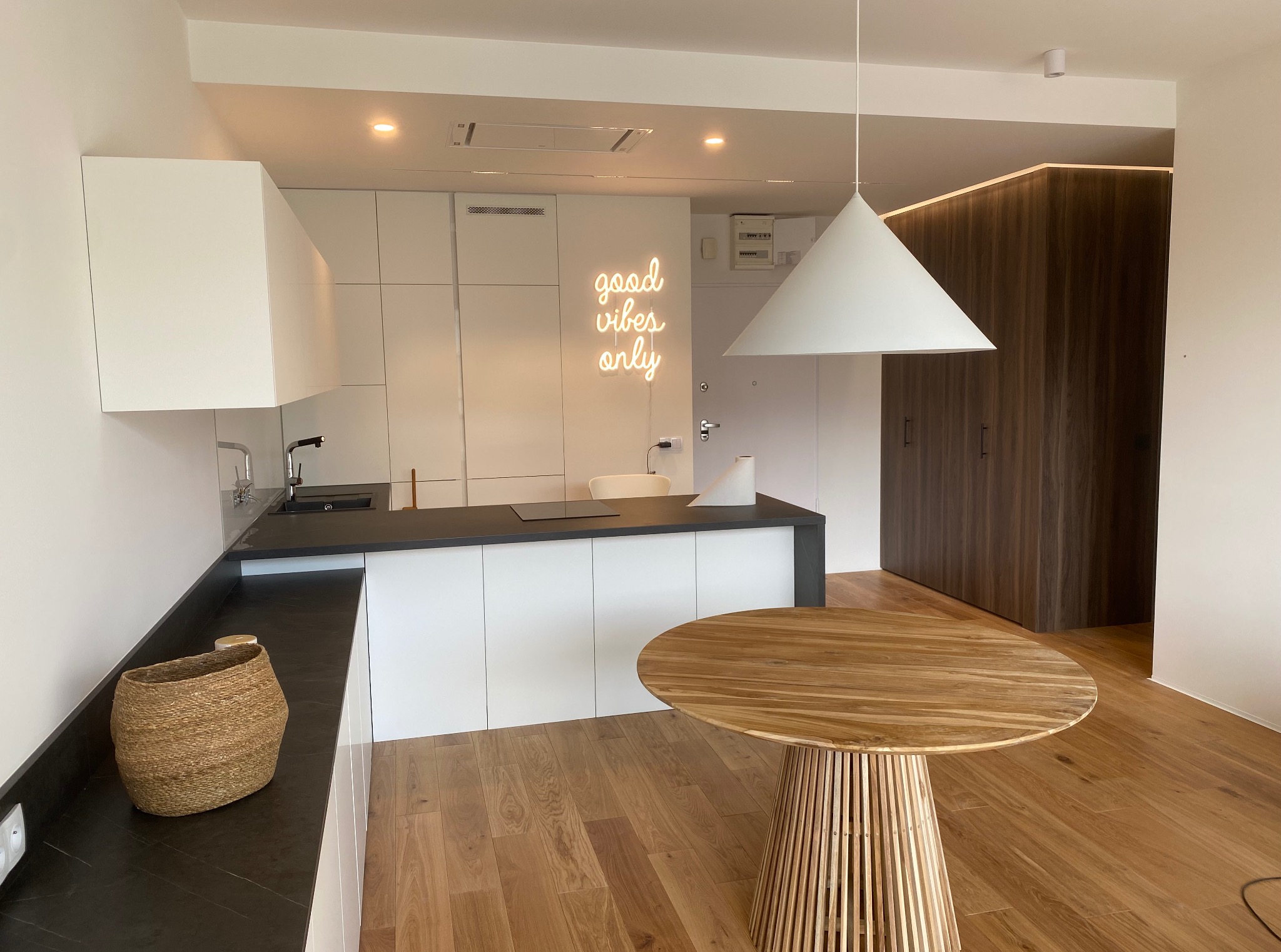 Nowoczesna, minimalistyczna kuchnia z białymi szafkami i czarnym blatem, oświetlona neonem 'good vibes only' i lampą stożkową. Na pierwszym planie drewniany stół.