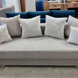 Jasnoszara sofa z poduszkami w showroomie meblowym, z widocznym fragmentem niebieskiego narożnika w tle. Ujęcie z przodu, naturalne oświetlenie.