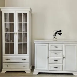 Biała przeszklona witryna z szufladami i komoda z dekoracją. Klasyczny zestaw mebli do salonu lub jadalni, z ciemnymi uchwytami i minimalistycznym wystrojem.