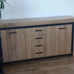 Drewniana komoda z szufladami i szafkami, czarne uchwyty, obok doniczka z rośliną, na podłodze panele. Minimalistyczny design, jasne wnętrze.